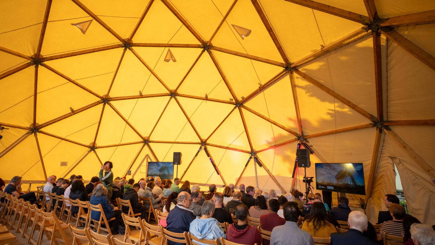people sat in chairs watching a talk under a dome shaped tent